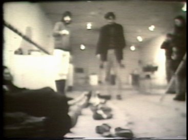 Still from Sequence 11 of Idea Demonstrations. Six spectators lie on the floor. Their shoes are removed and tied to a string which is then raised and dropped.